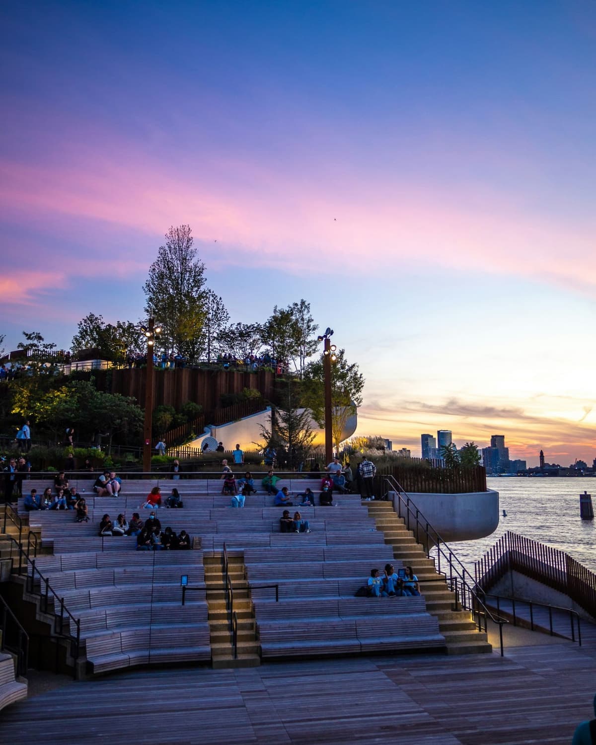 People sitting on tiered wooden seating at an outdoor amphitheatre at sunset, with the river and city skyline behind