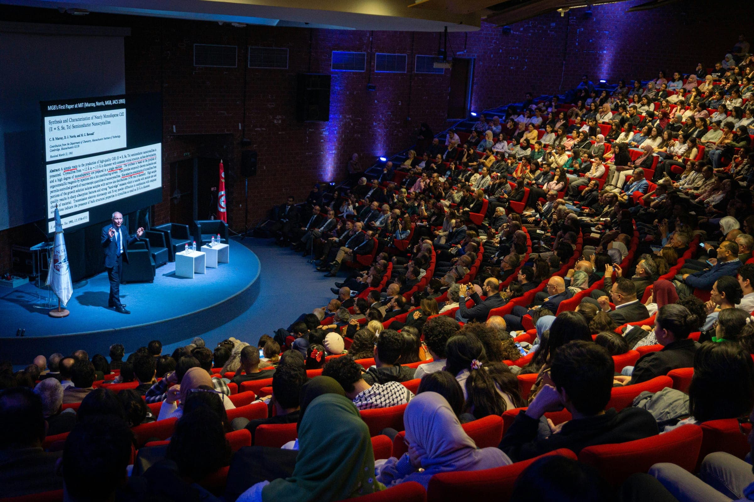 A speaker on stage addressing a full auditorium of seated attendees
