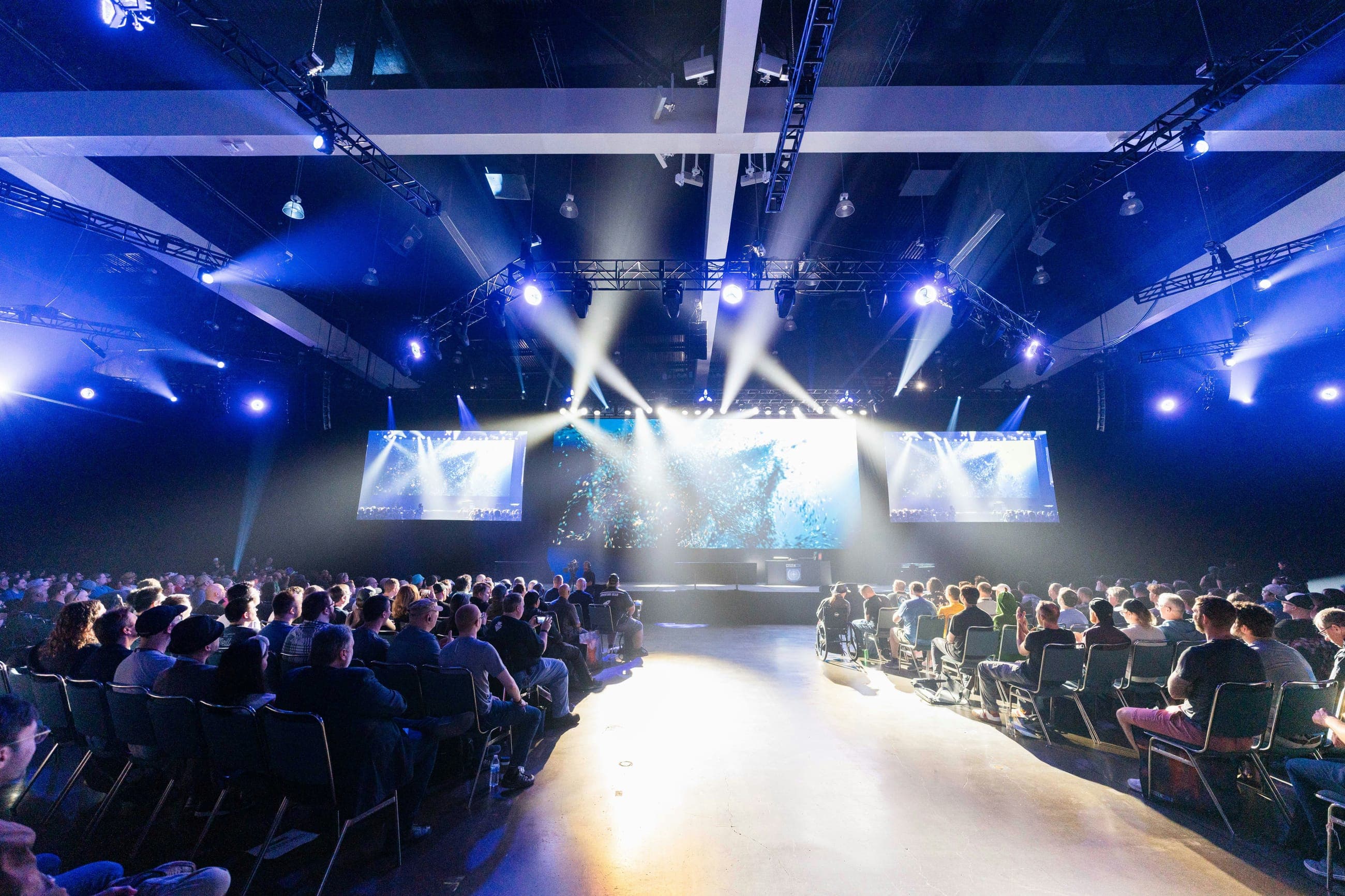 A packed auditorium watching a lit main stage with screens either side